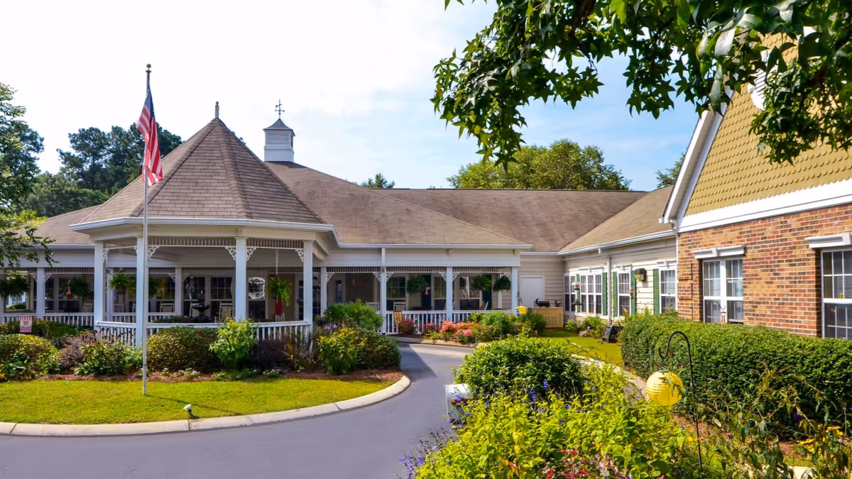 Exterior view of a senior living facility with a circular driveway, well-maintained garden beds, and a covered porch area. The building features a mix of brick and siding with multiple windows, and an American flag is displayed on a flagpole in the front lawn. Trees and greenery surround the facility under a partly cloudy sky.
