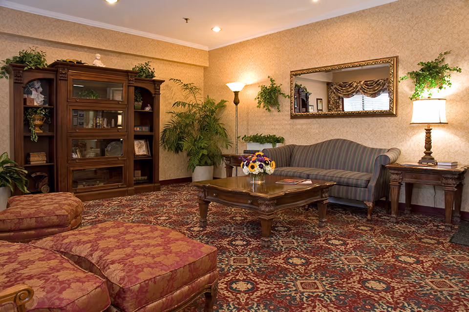 A cozy living room with a patterned carpet, a striped sofa, a wooden coffee table with a flower vase, two upholstered ottomans, a wooden cabinet with shelves holding decorative items, several potted plants, a floor lamp, a table lamp, and a large framed mirror on the wall.