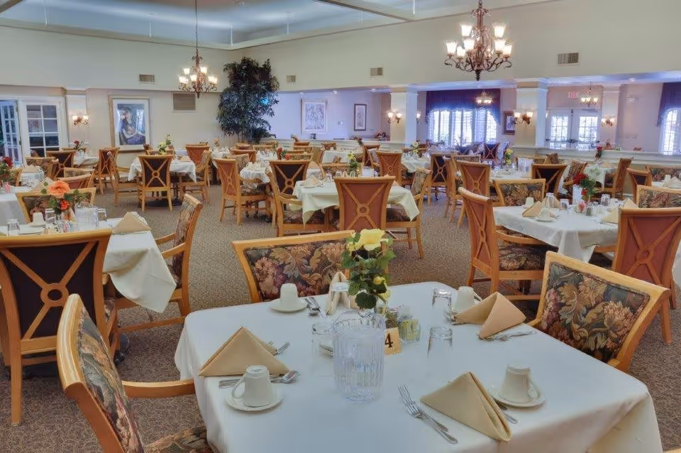 A spacious dining room with multiple tables covered in white tablecloths, each set with beige napkins, cups, glasses, and silverware. The chairs have wooden frames with floral upholstered backs and seats. The room is well-lit with chandeliers and wall sconces, and there are decorative plants and framed artwork on the walls.