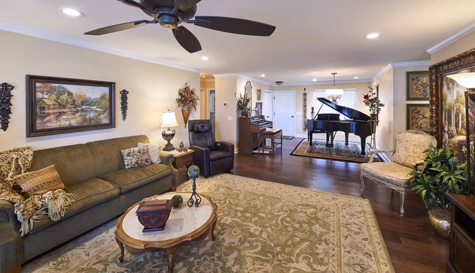 A spacious living room with a green sofa adorned with patterned pillows and a throw blanket, a wooden coffee table with decorative items, a recliner chair, and a grand piano on a rug near the entrance. The room features hardwood floors, a ceiling fan, framed artwork on the walls, and various plants and decorative pieces.