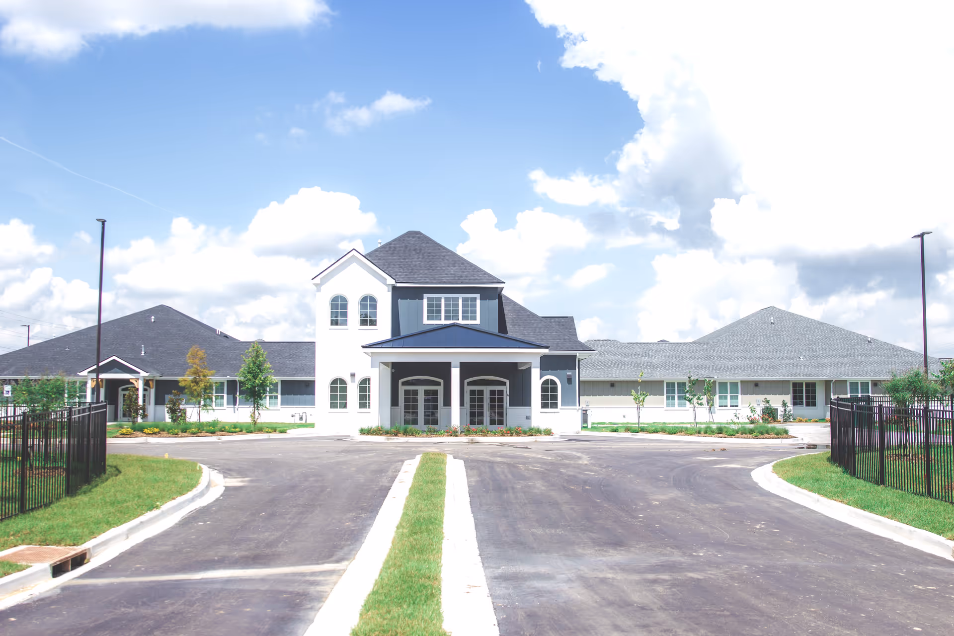 Front exterior view of a large, modern senior living facility building with a driveway leading up to the entrance, surrounded by green lawns and a partly cloudy blue sky.