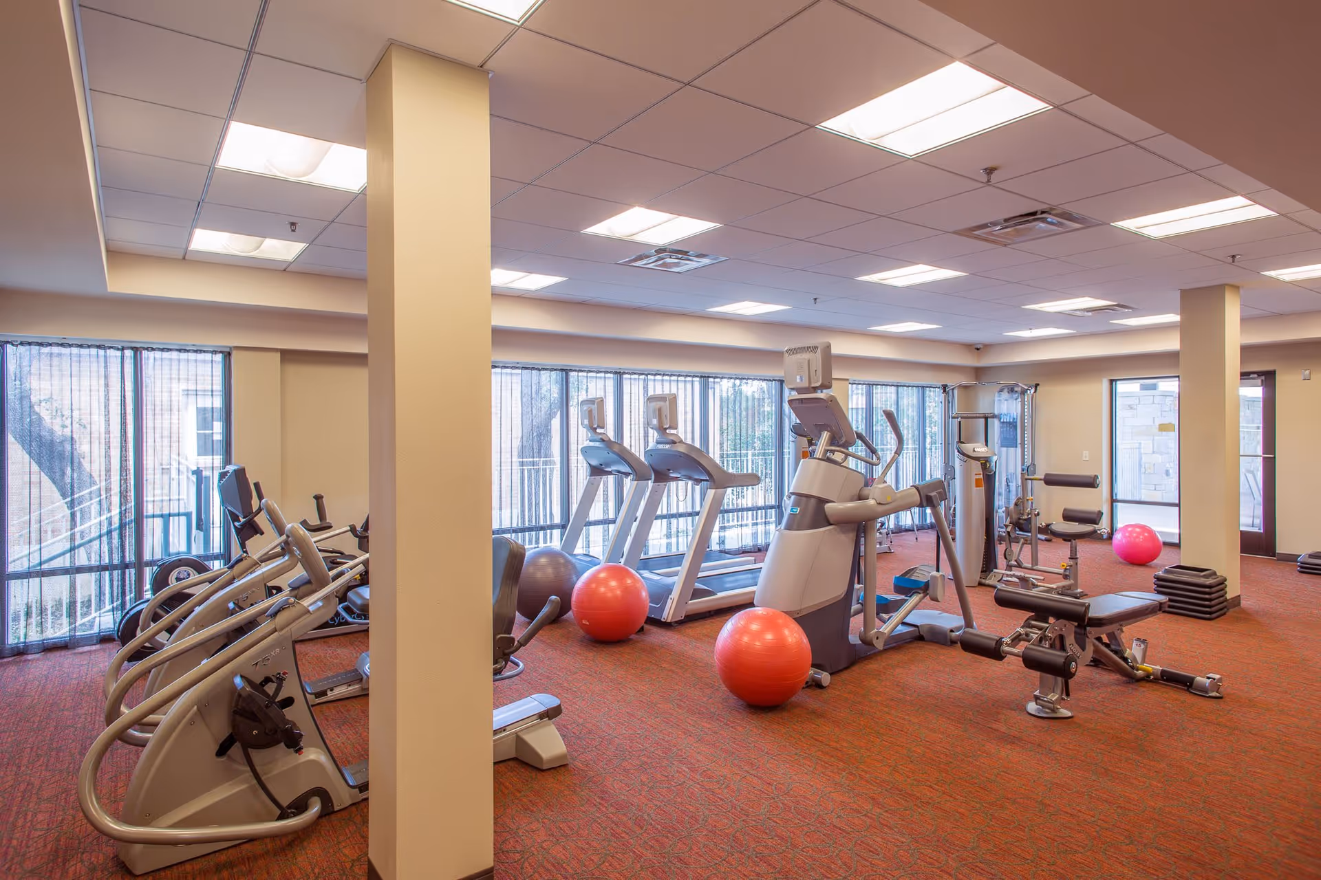 A well-lit indoor gym room with exercise equipment including treadmills, elliptical machines, weight machines, and red exercise balls. The room has large windows with sheer curtains and a red carpeted floor.