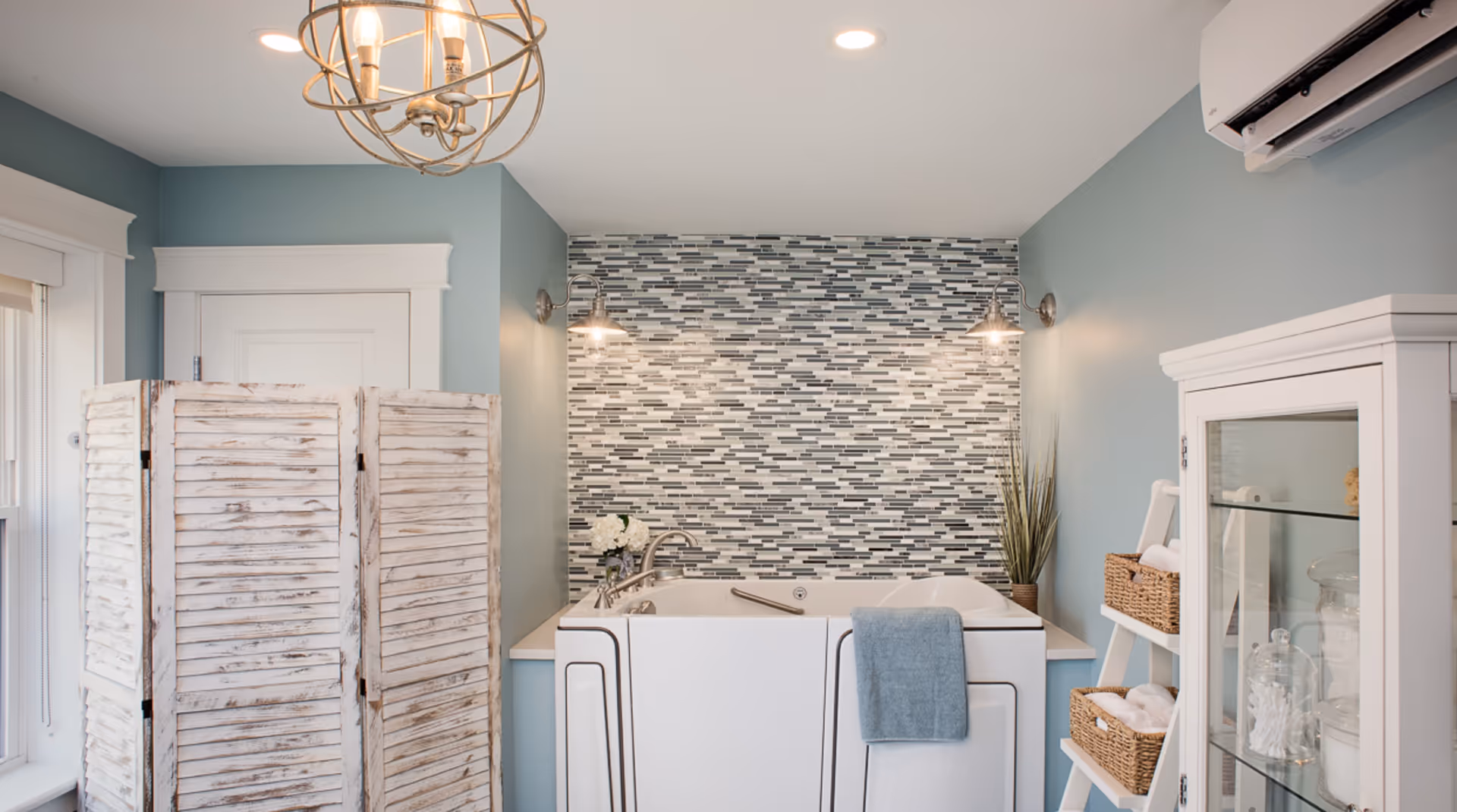 Bright bathroom with a walk-in tub against a gray mosaic tile accent wall, blue-green walls, a rustic folding screen, and white storage cabinet with baskets.