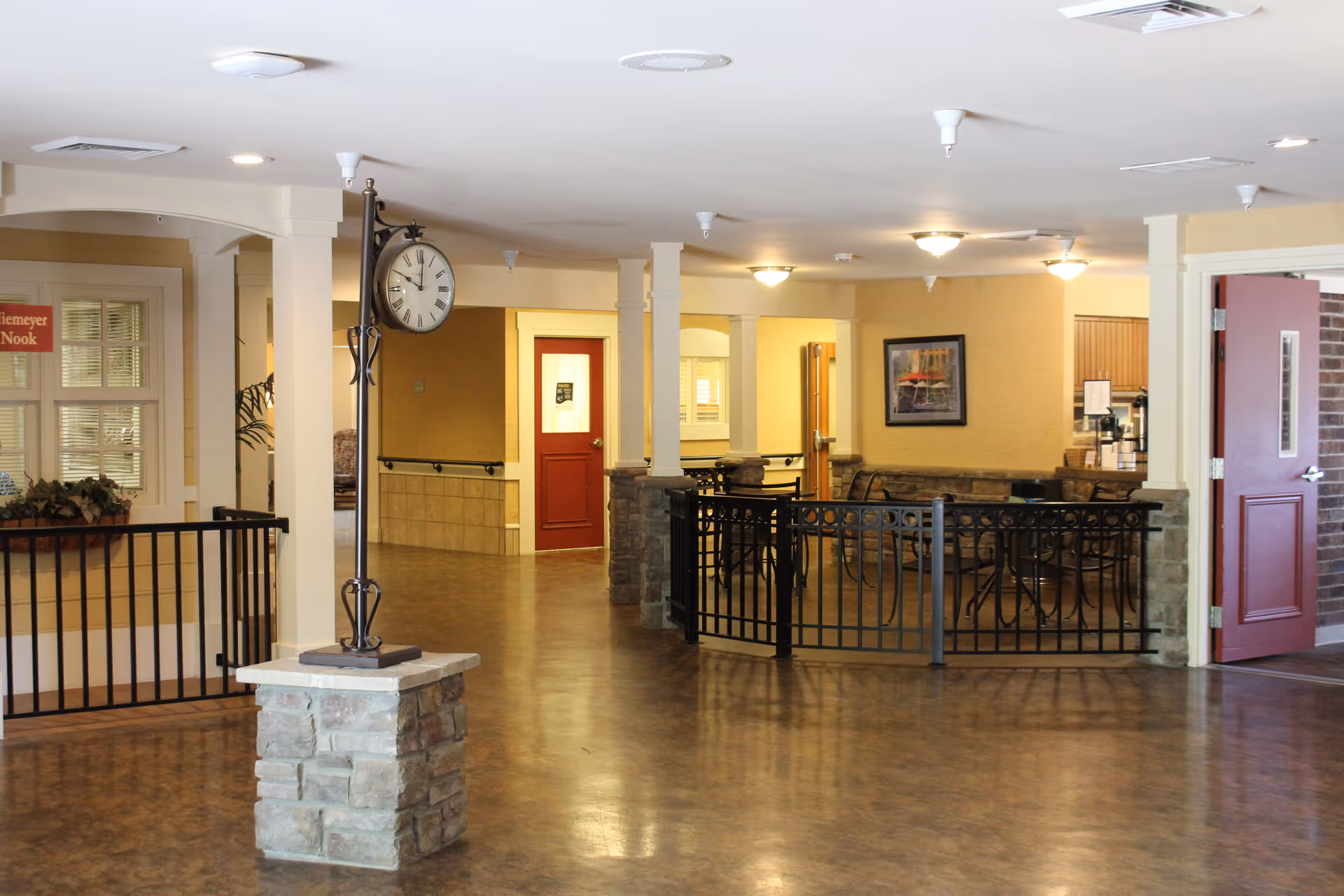 Interior view of a senior living facility hallway with a stone pillar supporting a vintage-style clock. The area has polished floors, cream-colored walls, and several doors including a red door. There is a seating area with tables and chairs behind a black metal railing. A framed picture hangs on the wall, and there is a sign that reads 'Siemeyer Nook' visible through a window on the left.