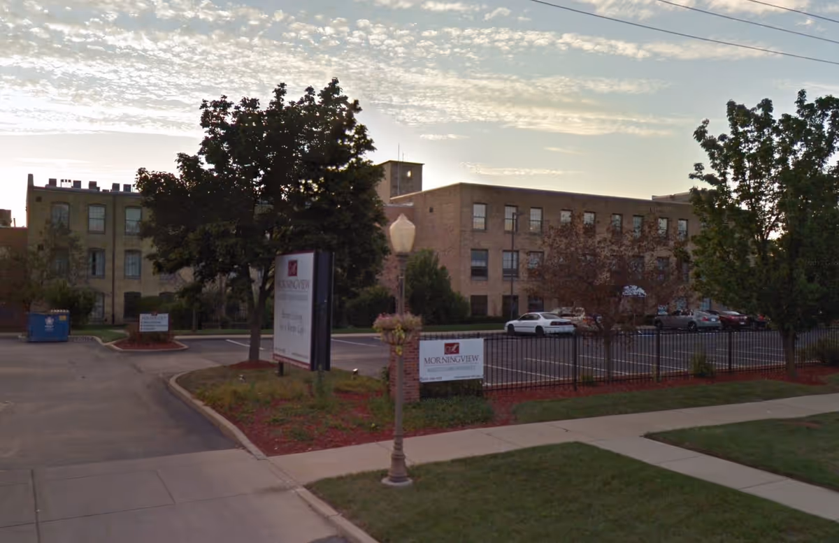 Exterior view of Morningview Assisted Living Residences showing a multi-story brick building with several windows, a parking lot with cars, trees, a sidewalk, and two signs displaying the facility's name near the entrance.