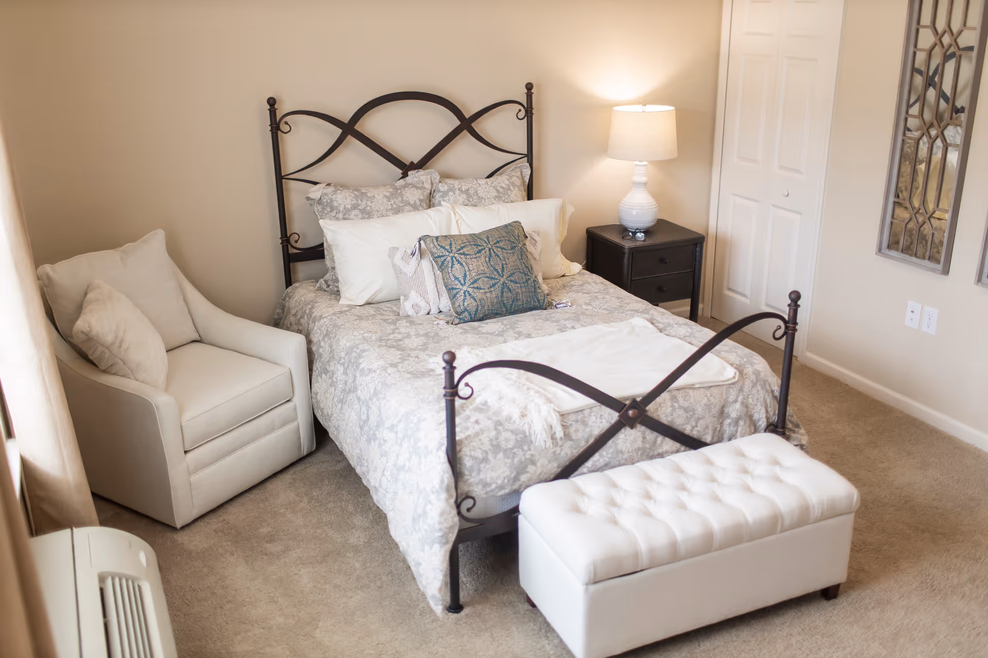 A cozy bedroom with a metal framed bed adorned with multiple pillows and a patterned bedspread. Next to the bed is a dark wooden nightstand with a white lamp on it. There is a cushioned armchair in the corner and a white tufted bench at the foot of the bed. The room has beige walls and carpeted floor, with two decorative mirrors hanging on the wall.