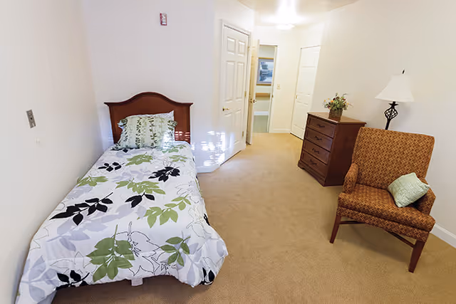 A simple bedroom in a senior living facility with a single bed covered in a white, green, and black leaf-patterned bedspread. There is a wooden headboard, a wooden dresser with a flower arrangement on top, a patterned armchair with a green pillow, and a floor lamp. The room has beige carpet and white walls, with doors leading to other areas in the background.