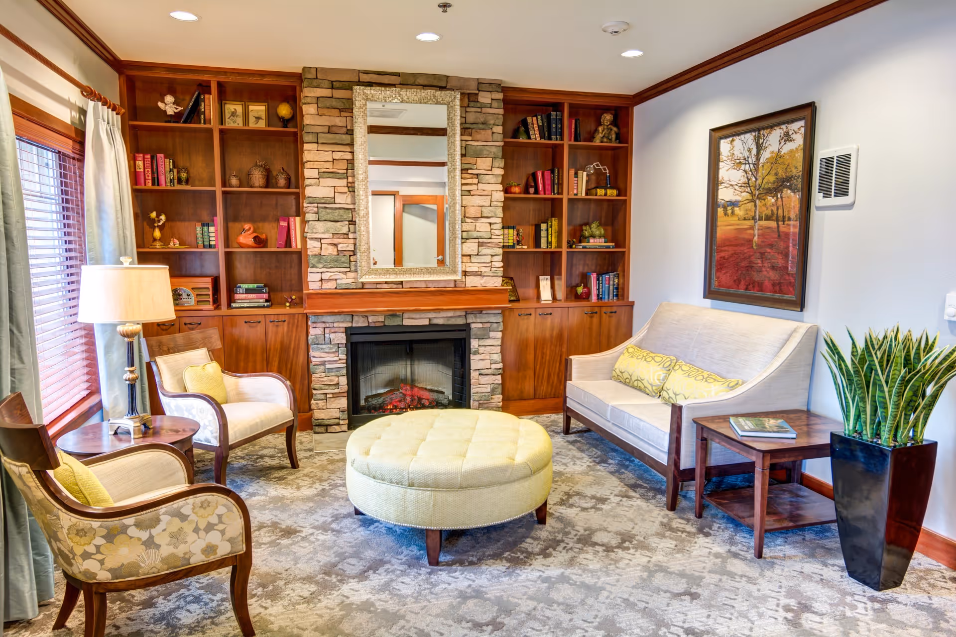 A cozy living room with a stone fireplace in the center, flanked by wooden built-in bookshelves filled with books and decorative items. In front of the fireplace is a round, tufted ottoman. To the left, there are two upholstered armchairs with floral patterns and a wooden side table with a lamp. To the right, there is a beige loveseat with two yellow patterned pillows, a wooden side table with a book, and a tall potted plant. A framed landscape painting hangs on the wall above the loveseat. The room has large windows with curtains and a patterned carpet.