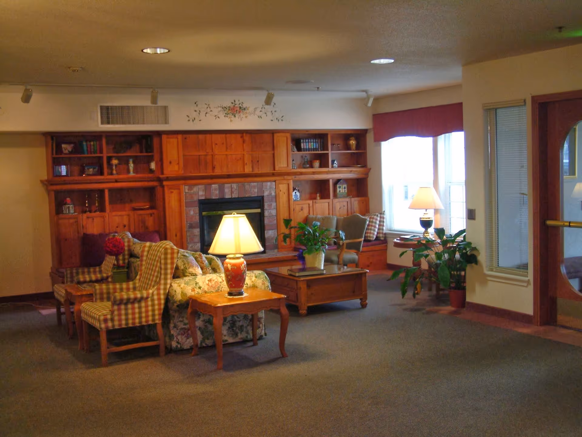 A cozy communal living room with upholstered chairs, a wooden built-in shelving unit and fireplace, table lamps, and potted plants.