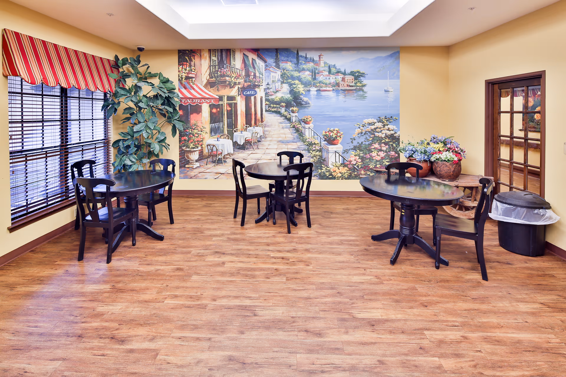 A dining area with three round dark wooden tables, each surrounded by black chairs. The room has wooden flooring, a large window with red and white striped valance, a large mural on the wall depicting a scenic outdoor café by a waterfront, and a corner table with flower pots and a trash can nearby.