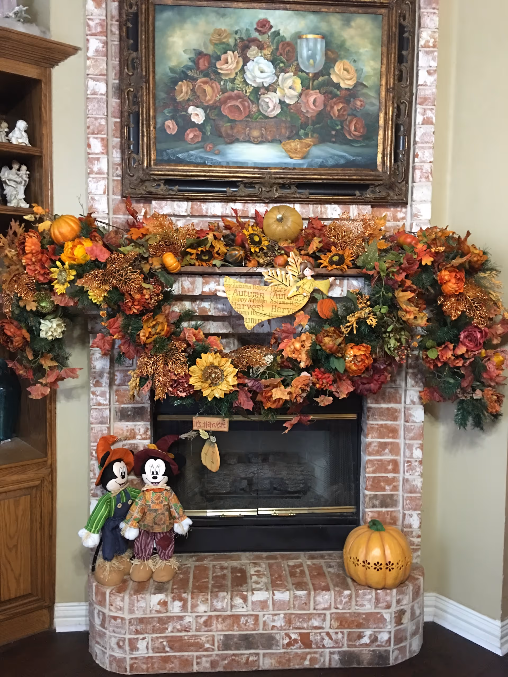 A brick fireplace decorated with autumn-themed garlands featuring orange, yellow, and red leaves, sunflowers, and small pumpkins. Two scarecrow dolls dressed in fall clothing stand on the hearth next to a large decorative pumpkin. Above the fireplace hangs a framed painting of a floral arrangement with roses and a goblet. To the left is a wooden cabinet with white figurines displayed.