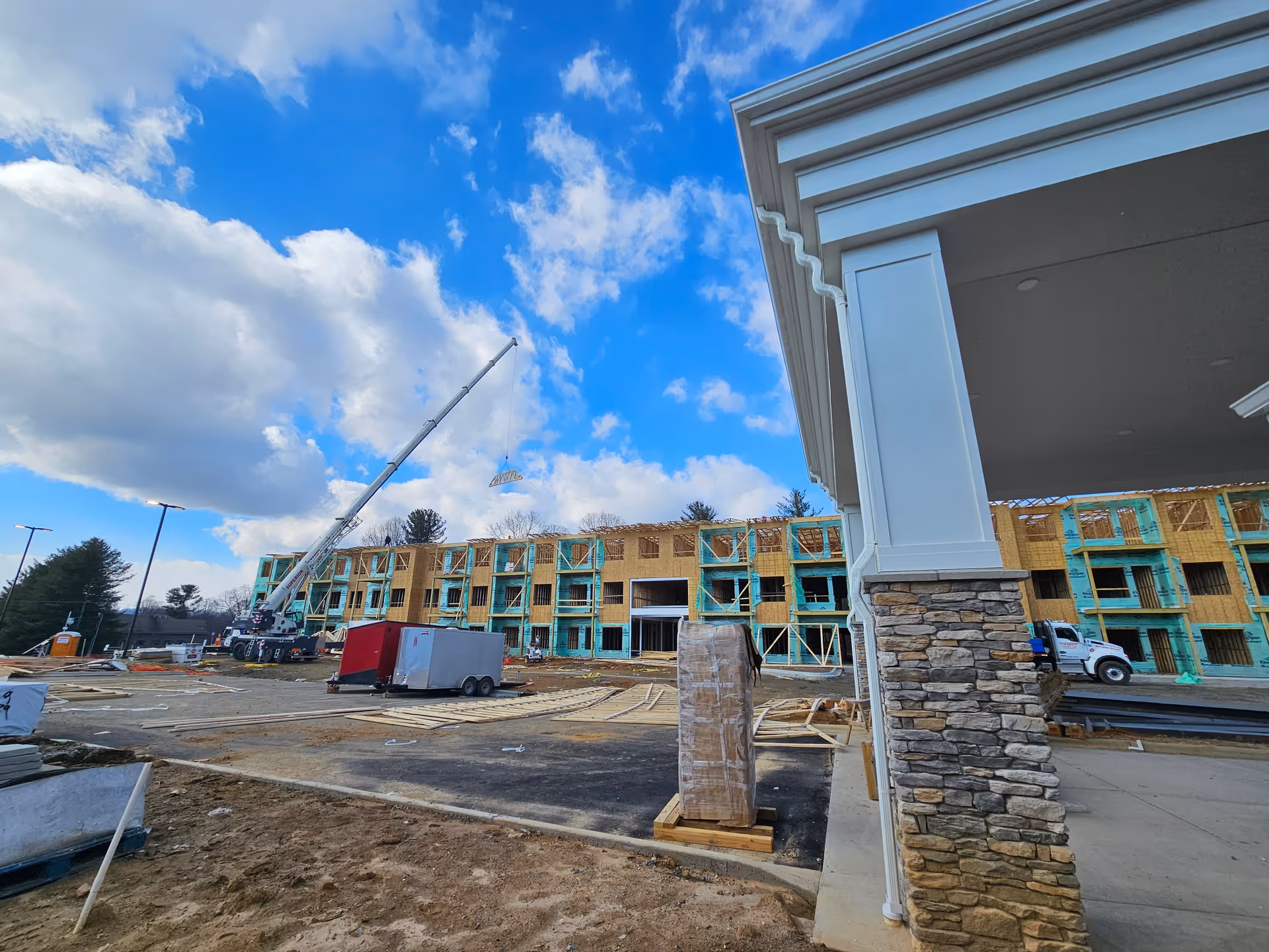 Construction site of a multi-story building under a blue sky with scattered clouds. A crane is lifting materials to the upper floors. The building framework is visible with some areas covered in green insulation. In the foreground, there is a stone and white column structure, construction trailers, and construction materials scattered around the site.