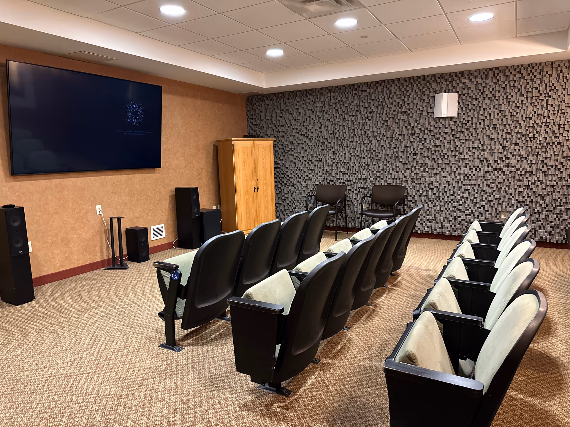A small theater room with two rows of theater-style seating facing a large flat screen TV mounted on a brown wall. The room has a textured gray acoustic wall panel on one side, two black chairs against that wall, and a wooden cabinet near the TV. The floor is carpeted and the ceiling has recessed lighting.
