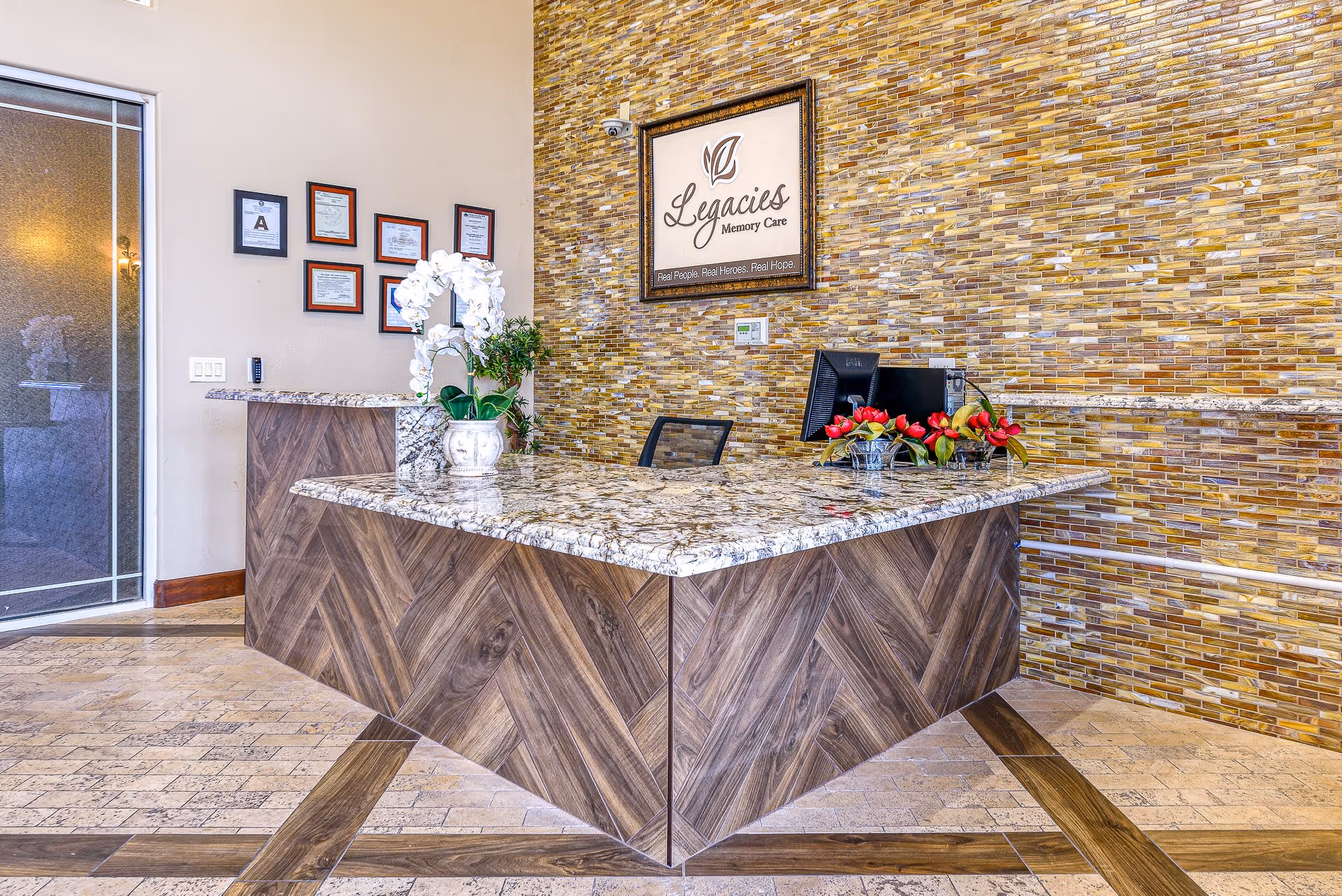 Reception desk area with a granite countertop and wood paneling. Behind the desk is a wall with yellow and brown mosaic tiles and a framed sign that reads 'Legacies Memory Care Real People. Real Heroes. Real Hope.' There are several framed certificates on the adjacent wall and decorative plants on the desk.