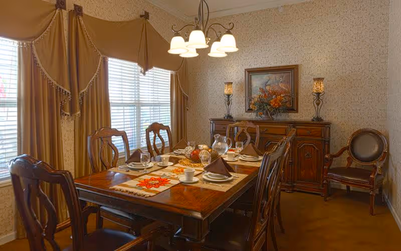 A traditional dining room with a wooden dining table set for six people, featuring plates, cups, napkins, and a centerpiece. The room has patterned wallpaper, a wooden sideboard with decorative items and a framed painting above it, a chandelier with five lights hanging from the ceiling, and large windows with beige curtains.