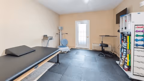A small therapy or exercise room with a black padded treatment table, exercise equipment including a vibration plate, resistance bands, dumbbells, and a stool. The room has beige walls, a door with a window, and black interlocking foam floor mats.