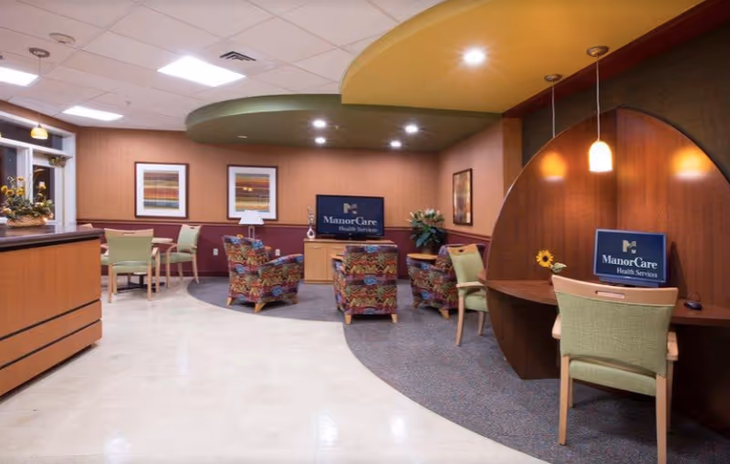 Interior view of a senior living facility common area with patterned armchairs, green chairs, a reception desk, and two computer stations with ManorCare Health Services logos on the screens. The room has warm lighting, framed artwork on the walls, and a mix of carpet and tile flooring.