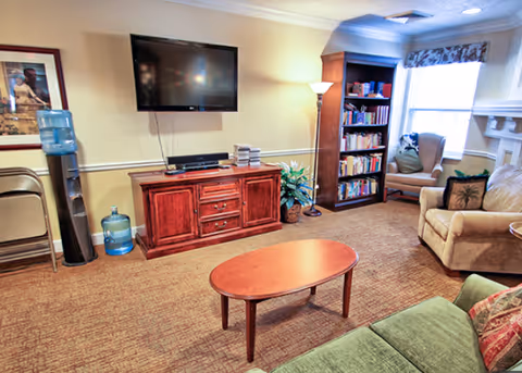 A cozy living room with a green sofa, a beige armchair, a wooden coffee table, a wooden TV stand with a flat-screen TV mounted above it, a bookshelf filled with books, a floor lamp, a water cooler, and a window with floral curtains letting in natural light.