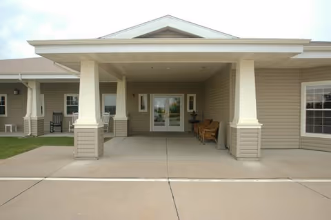 Entrance of Windsor Manor Assisted Living Community showing a covered driveway with two large white columns, a concrete walkway, and double glass doors. There are benches and rocking chairs on the porch area beside the entrance.