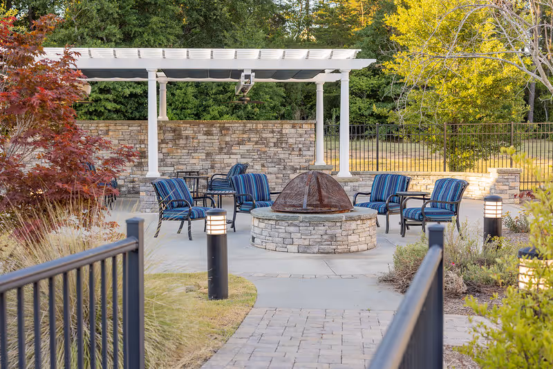 Outdoor courtyard with a central stone fire pit surrounded by striped chairs beneath a white pergola, paved walkways and landscaping.
