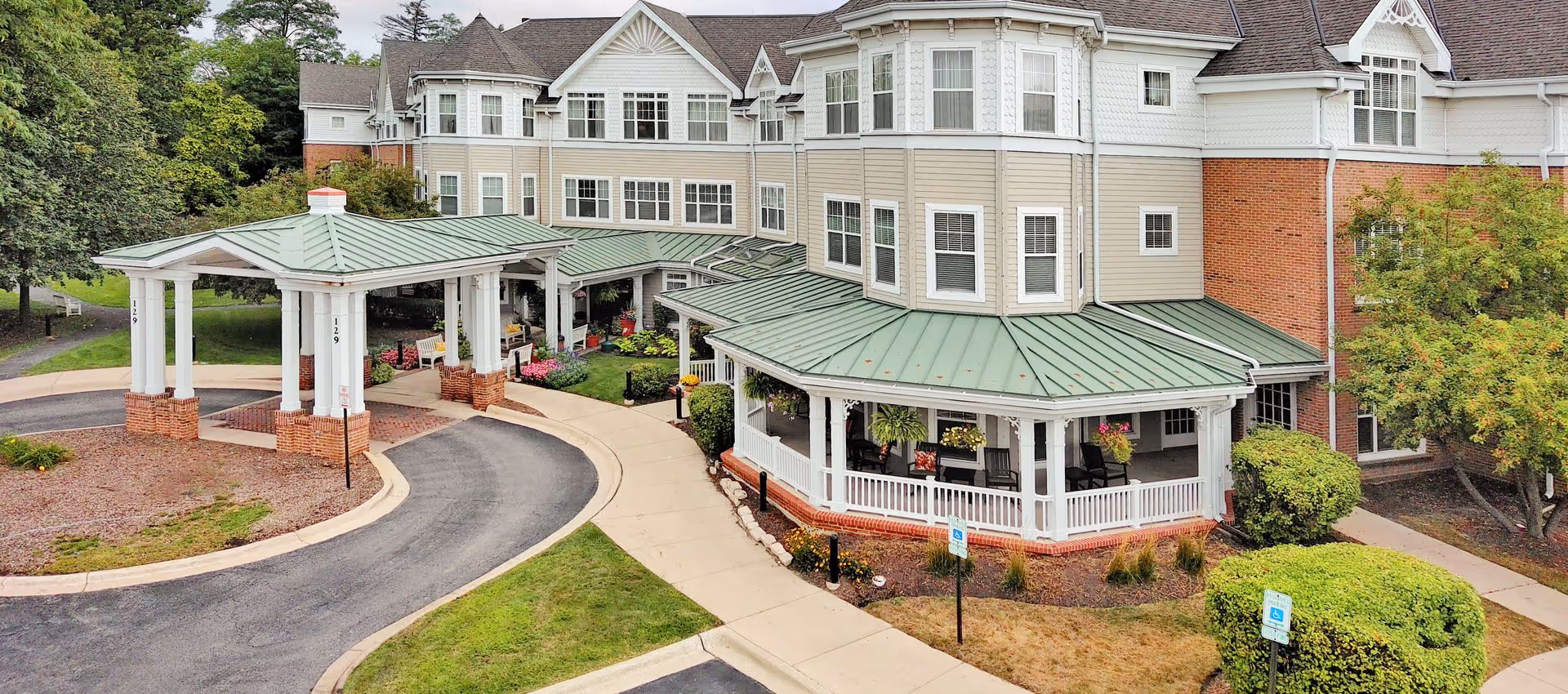 Exterior view of a senior living facility with a covered entrance and porch area. The building has multiple windows, beige siding with white trim, and a green metal roof. There are landscaped bushes, trees, and flower beds around the entrance and along the sidewalk.