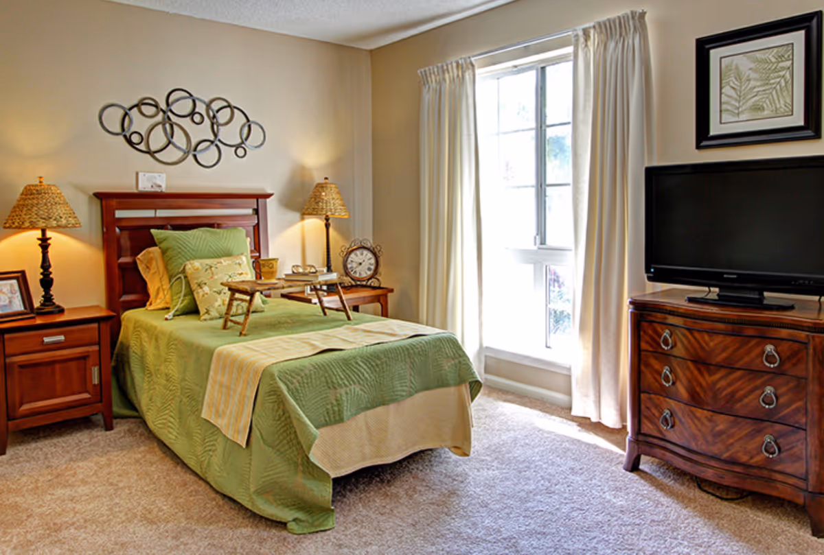 Sunlit bedroom with a twin bed in green bedding, wooden nightstands, decorative wall art, and a TV on a dresser.