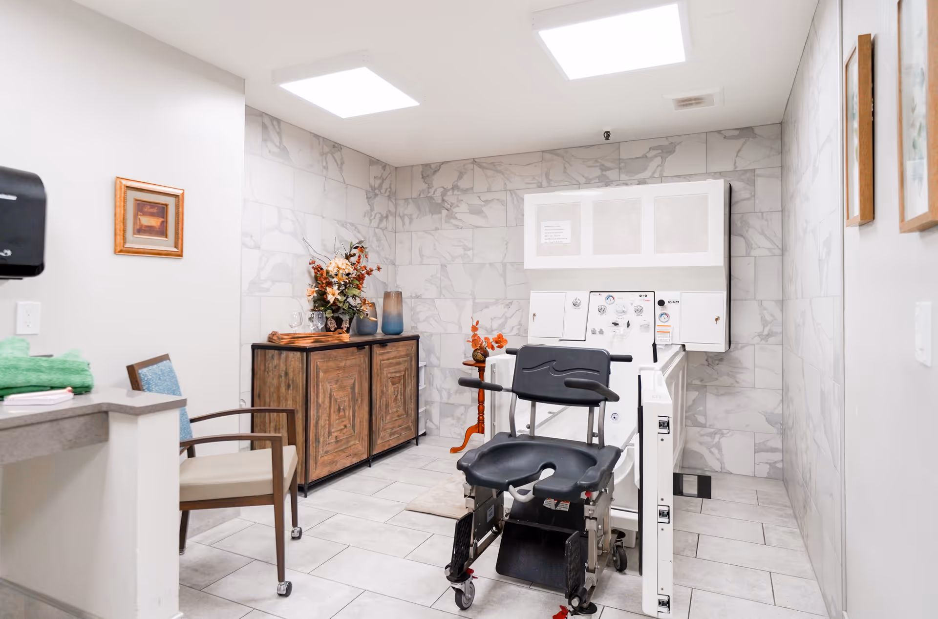 A clean and well-lit room with marble-patterned walls and tiled floor, featuring a medical bathing chair and equipment, a wooden cabinet with decorative flowers, a chair with armrests, and framed artwork on the walls.