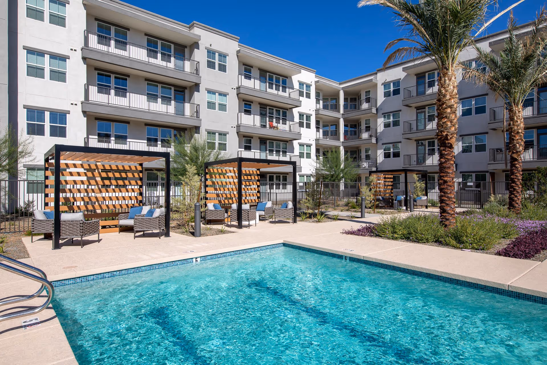Outdoor swimming pool area with clear blue water, surrounded by a concrete deck. There are three shaded seating areas with modern wicker chairs and blue cushions. The background features a multi-story residential building with balconies and large windows, along with palm trees and landscaped greenery under a clear blue sky.