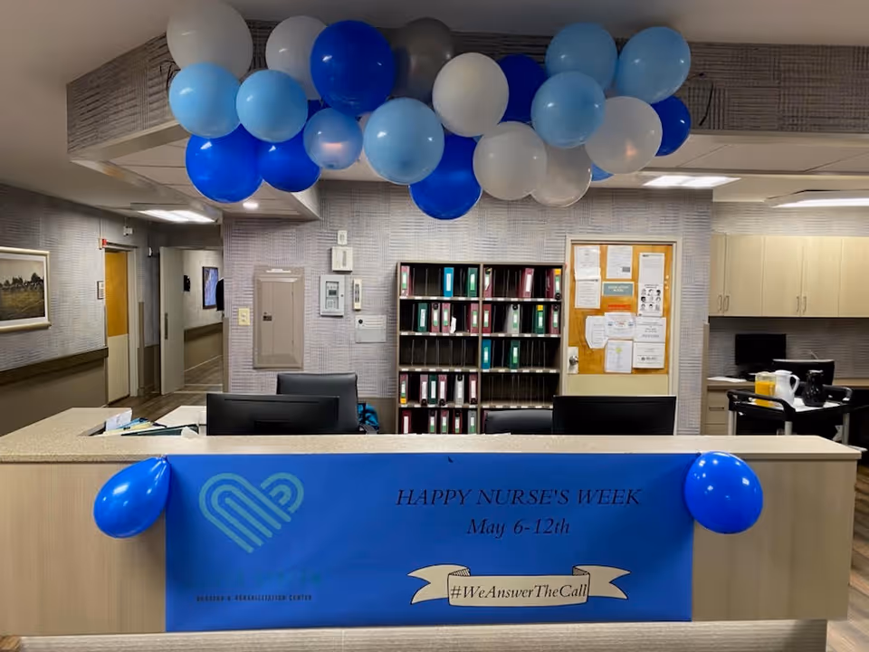 Reception desk area decorated with blue, white, and gray balloons hanging from the ceiling and attached to the front of the desk. A blue banner on the desk reads 'HAPPY NURSE'S WEEK May 6-12th #WeAnswerTheCall'. Behind the desk are shelves with binders and a bulletin board with various notices. The room has neutral-colored walls and cabinets, with a hallway visible to the left.