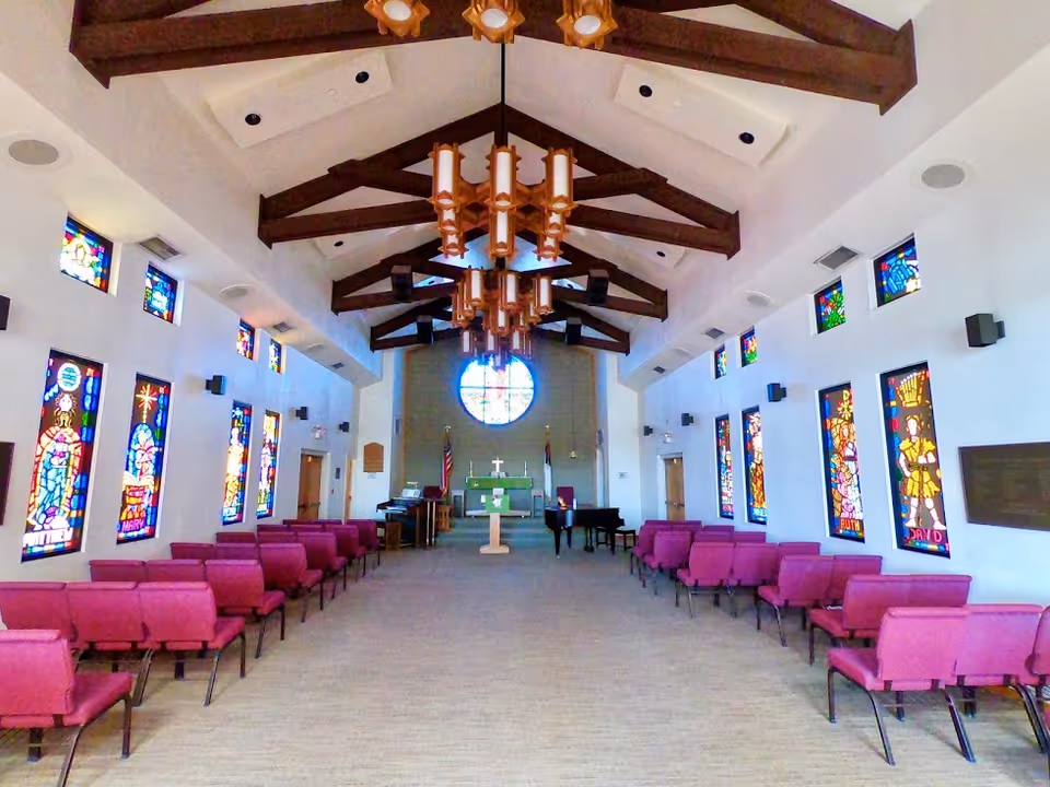 Interior view of a chapel with rows of purple chairs on both sides, stained glass windows along the walls, wooden beams on the ceiling, and a podium with a cross at the front near an altar and piano.
