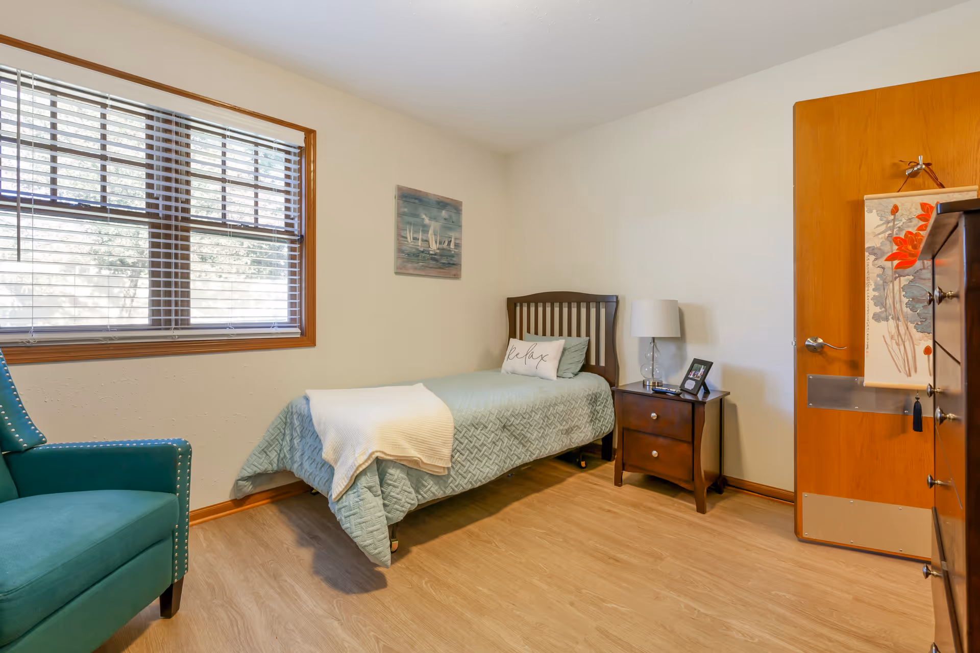 A small bedroom with a single bed covered in a light blue quilt and a white blanket. There is a pillow with the word 'relax' on it. Next to the bed is a wooden nightstand with a glass lamp and a framed photo. A teal armchair is positioned near a window with white blinds. The room has light-colored walls, wood flooring, and a wooden door with a decorative hanging.