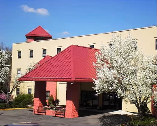 Exterior view of a senior living facility building with a beige facade and a red metal roof over the entrance. There are blooming white flowering trees near the entrance and a clear blue sky in the background.
