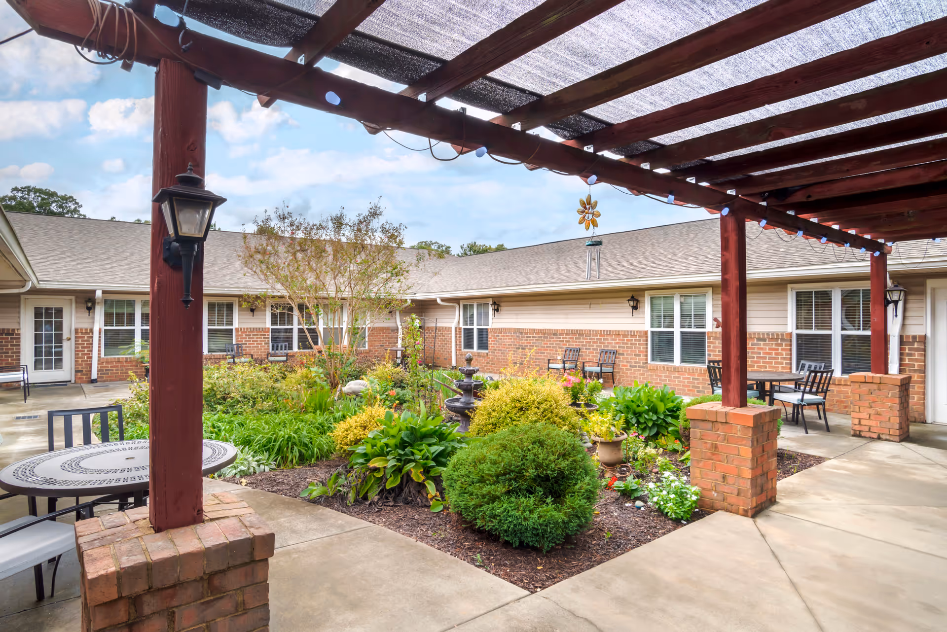 Outdoor courtyard area at Brookdale Monroe Square featuring a garden with various green plants and flowers, a central water fountain, and seating areas with tables and chairs under a wooden pergola with a mesh shade cover. The courtyard is surrounded by a single-story building with brick and siding exterior walls and multiple windows and doors.