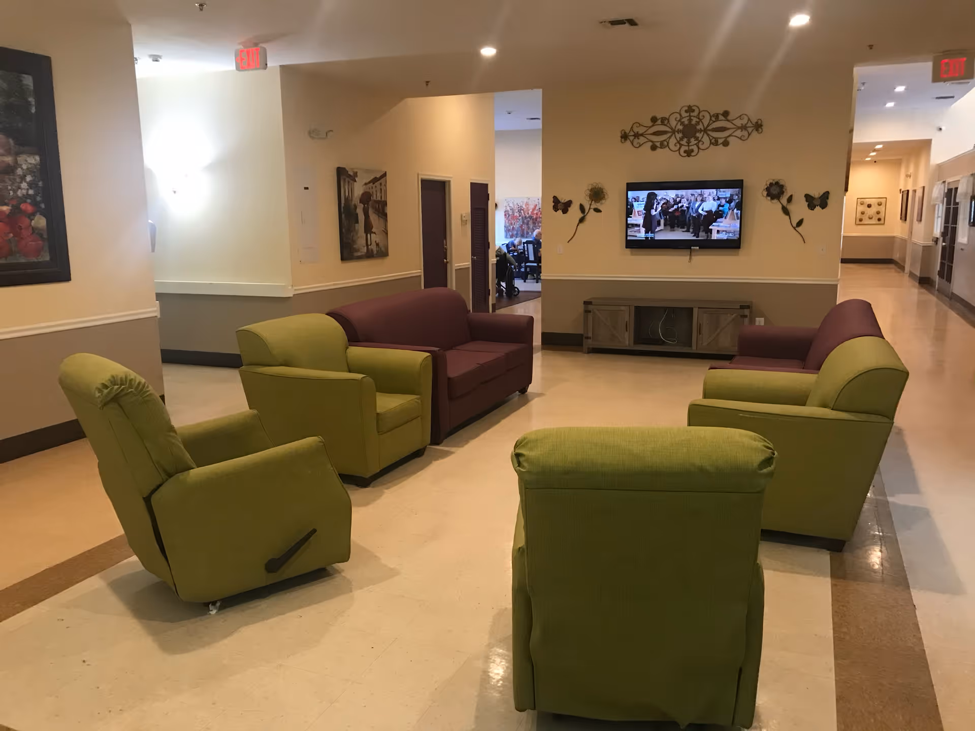 A common area in a senior living facility with two green armchairs, two green recliners, and two purple sofas arranged in a circle facing a wall-mounted TV. The walls are decorated with artwork and floral wall hangings. The space is well-lit with overhead lighting and has a beige and brown tiled floor.