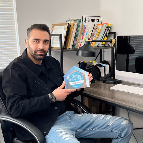 A man with dark hair and beard sits in an office chair at a desk holding a blue and white award plaque that reads 'Best of Senior Living 2023 Winner'. Behind him is a bookshelf with books and a small sign that says 'OFFICE'. On the desk are a computer monitor, keyboard, desk lamp, and office supplies.
