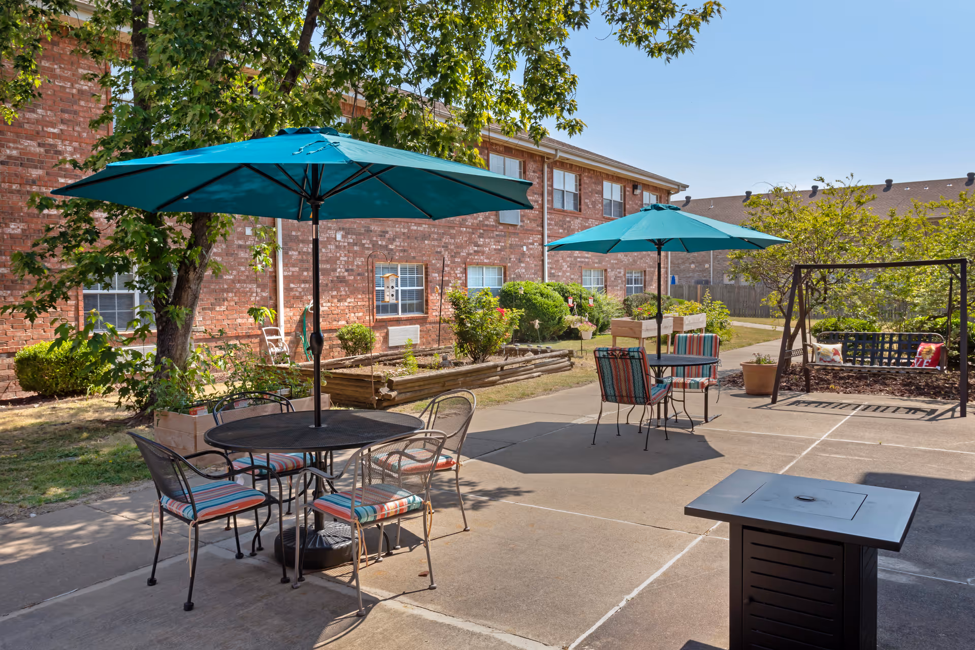 Outdoor patio area with two round metal tables, each shaded by a teal umbrella. Each table is surrounded by metal chairs with striped cushions. There is a brick building with windows and greenery in the background. A wooden swing with cushions is visible on the right side, along with a fire pit in the foreground.