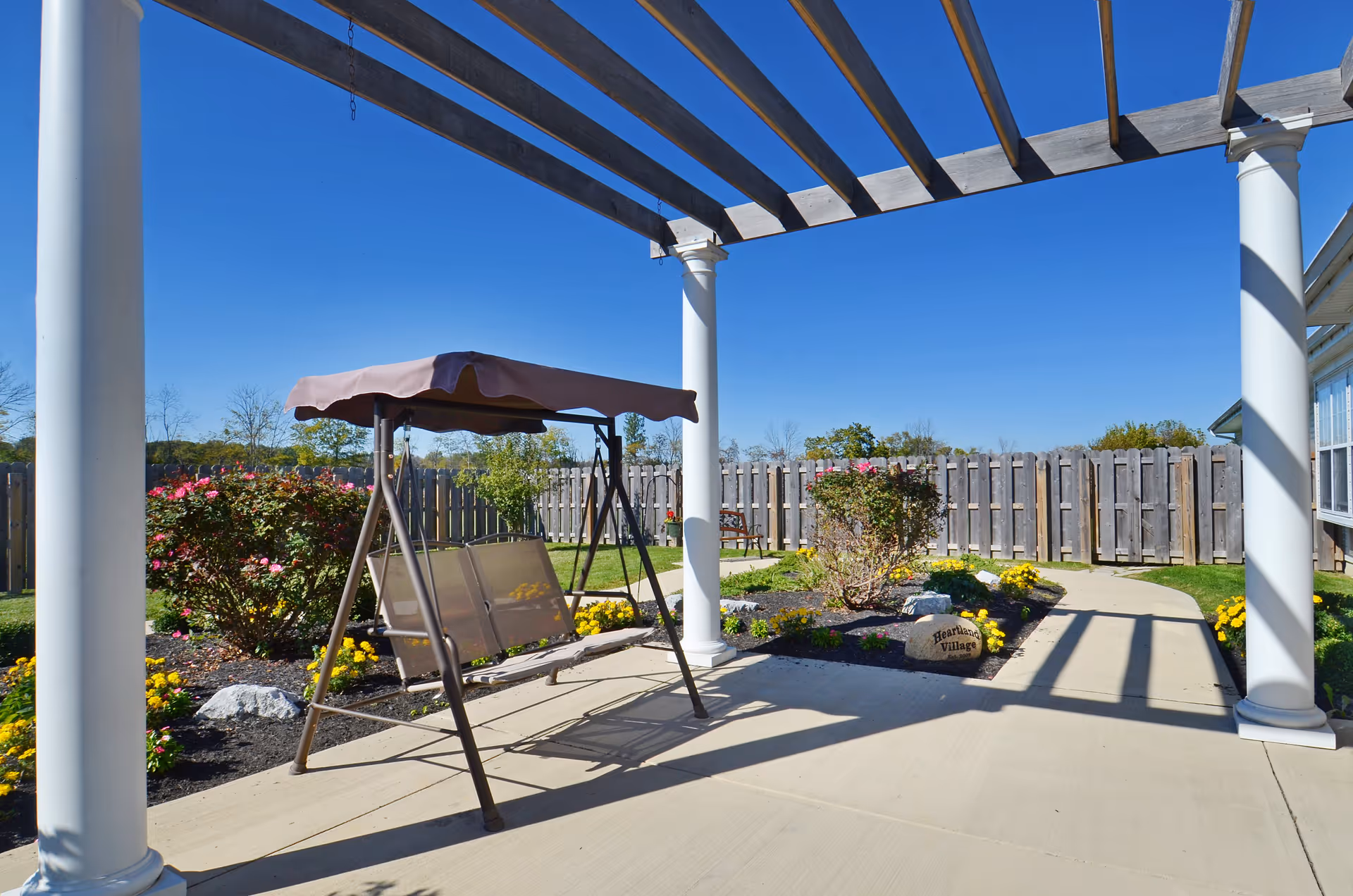 Outdoor patio area with a swing bench under a pergola, surrounded by flower beds with blooming flowers and shrubs, a wooden fence in the background, and a clear blue sky.
