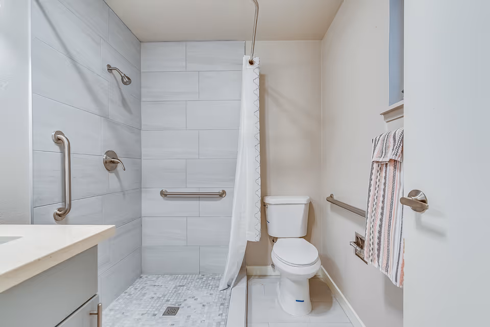 A clean and modern bathroom featuring a walk-in shower with gray tiles and stainless steel grab bars, a white toilet, a towel rack with a striped towel, and a white countertop with a sink partially visible on the left.