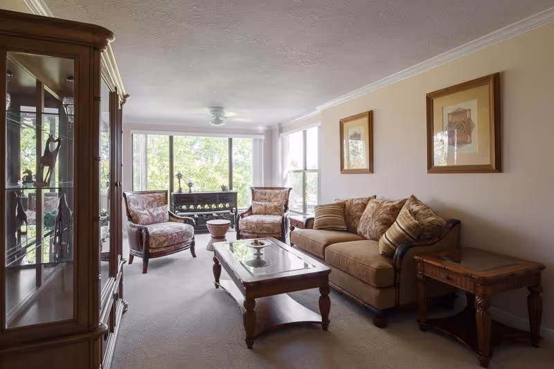 A cozy living room with a brown sofa adorned with patterned cushions, two matching armchairs with floral upholstery, a wooden coffee table with a glass top, and a wooden side table. The room has large windows letting in natural light and a wooden display cabinet on the left side.