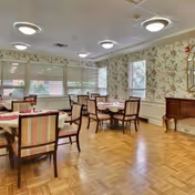 A dining room with several tables and chairs arranged neatly. The room has floral wallpaper, large windows with blinds, and a wooden floor. There is a wooden sideboard against one wall and ceiling lights providing illumination.