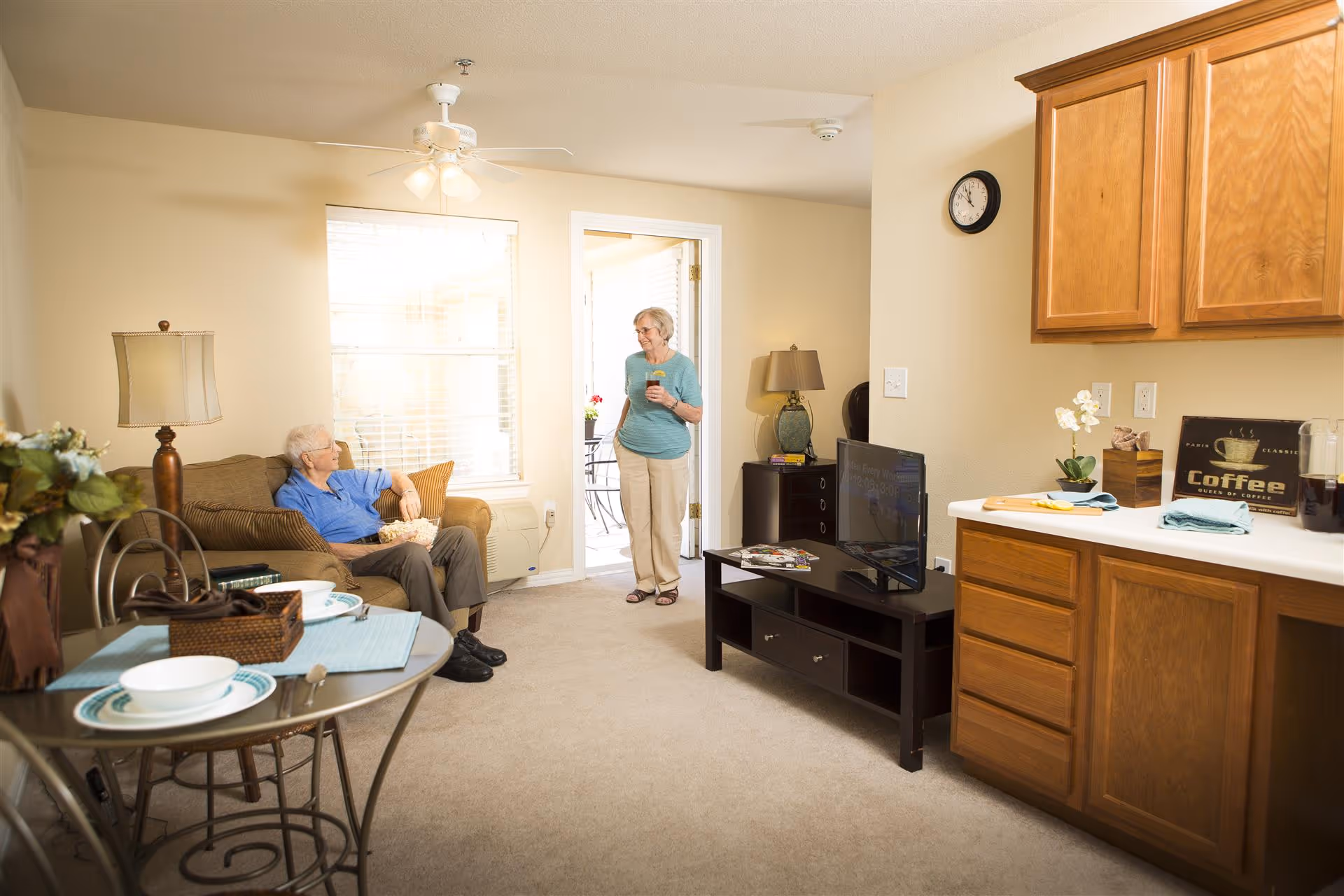 A cozy living room area in an assisted living facility with an elderly man sitting on a brown couch holding a bowl of popcorn, and an elderly woman standing near a doorway holding a drink. The room features beige walls, a ceiling fan with lights, a small round dining table set for two, a TV on a dark wooden stand, and wooden kitchen cabinets with a coffee sign on the counter.
