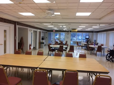 Interior view of a large room with multiple tables and chairs arranged for group seating. The room has a tiled ceiling with fluorescent lighting, large windows along one side, and a few people seated at the tables. There is a wheelchair visible on the right side and a television mounted on a stand near the windows.