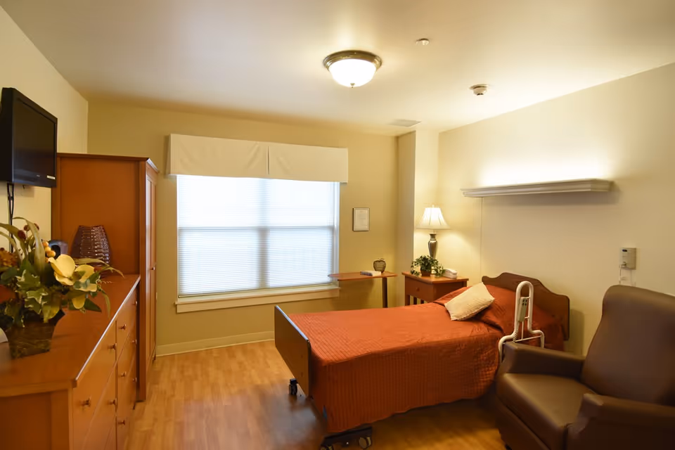 A cozy bedroom in a senior living facility featuring a single bed with an orange bedspread, a brown armchair, a wooden dresser with a flower arrangement, a wall-mounted TV, a window with blinds and a valance, a small side table with a lamp, and light-colored walls and flooring.