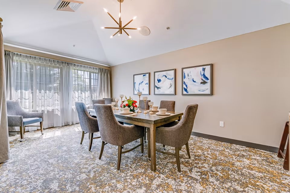 A bright dining room with a rectangular wooden table set for six people with beige plates and decorative goblets. The room features six upholstered chairs around the table, a patterned carpet, three abstract paintings on the beige wall, and large windows with sheer curtains allowing natural light to fill the space.