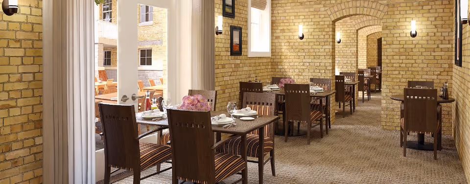 Dining area with multiple tables and chairs set for a meal, featuring brick walls, arched doorways, and soft lighting. There is a glass door leading to an outdoor seating area with wicker chairs.