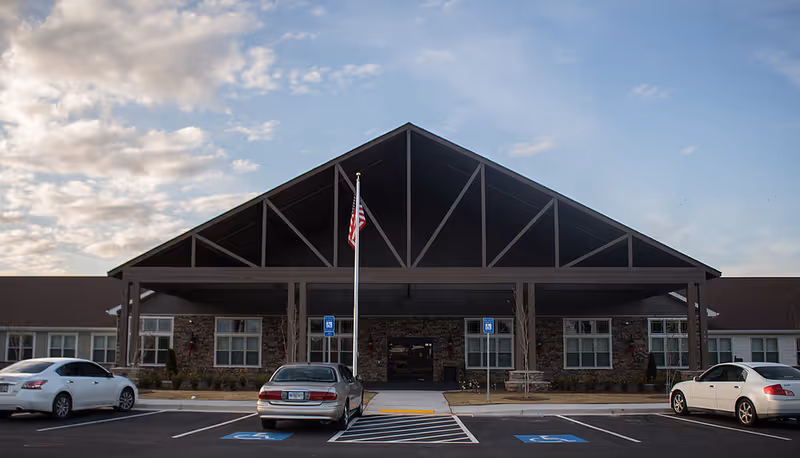 Front exterior view of The Phoenix at Lake Joy facility showing a large covered entrance with a peaked roof, an American flag on a flagpole, several parked cars, and a clear sky with some clouds.