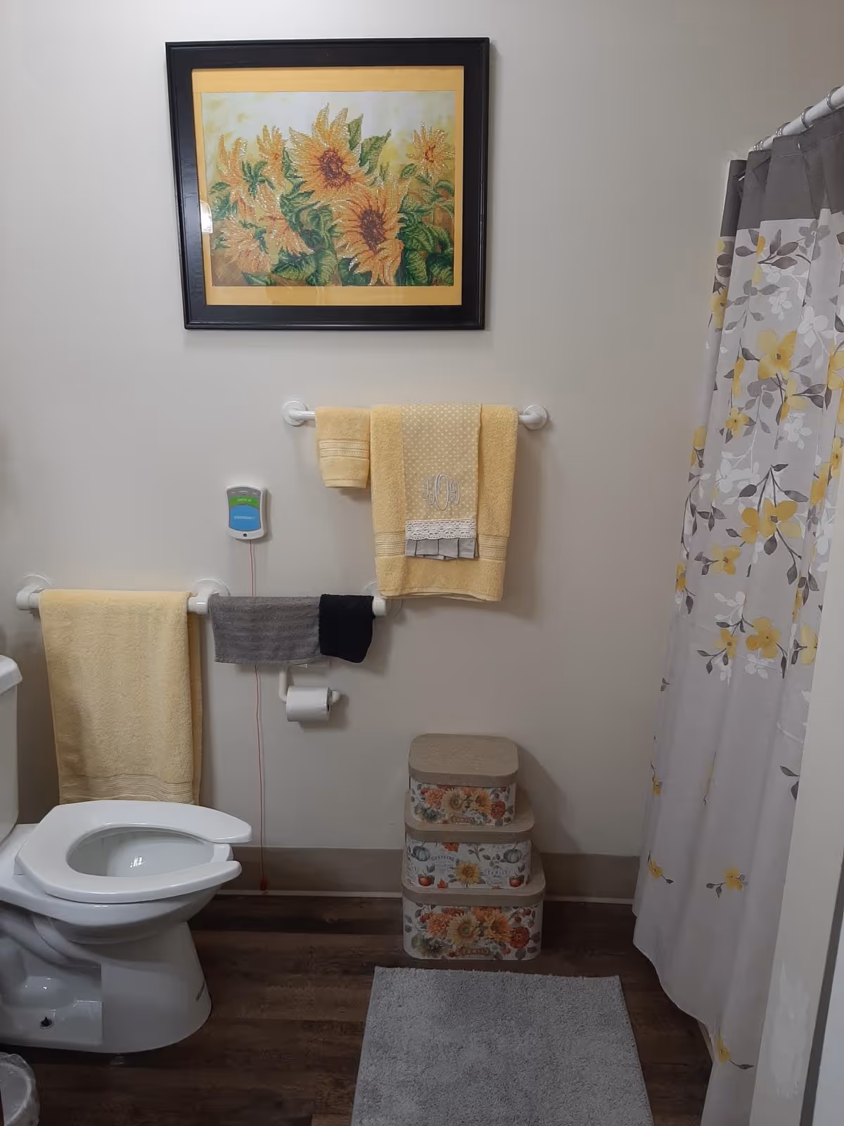 A bathroom with a white toilet, yellow and gray towels hanging on white towel racks, a floral shower curtain with yellow and gray flowers, a gray bath mat on a wooden floor, three stacked floral storage boxes, and a framed painting of sunflowers on the wall.