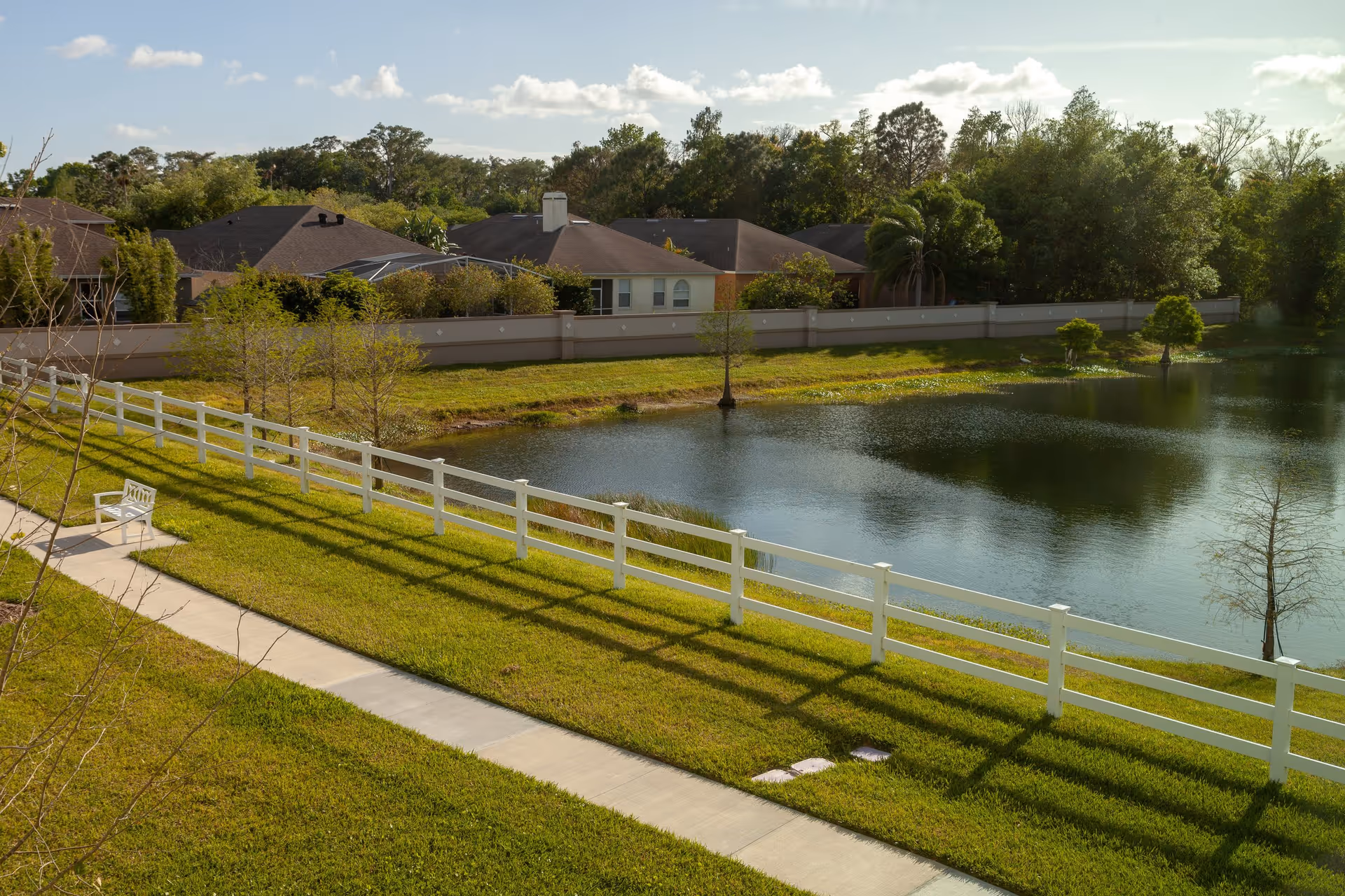 Sidewalk and white fence along a grassy lawn beside a small pond with a bench, with houses and trees in the background.