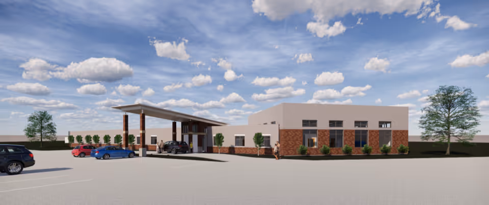 Exterior view of a single-story senior living facility building with a covered entrance. Several cars are parked in the parking lot in front of the building. The sky is partly cloudy with scattered white clouds. There are small trees and shrubs planted around the building.
