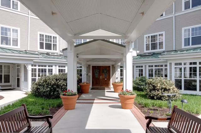 Covered entrance walkway leading to double wooden doors of a senior living facility, flanked by large potted plants and benches on either side, with windows and greenery visible on the building exterior.
